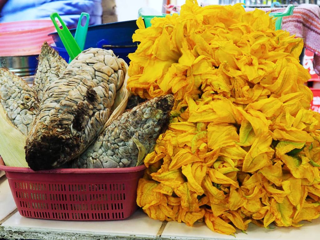 Huitlacoche, a gray corn fungus with swollen kernels, in a red basket next to a pile of yellow squash blossoms, two weird Mexican foods.