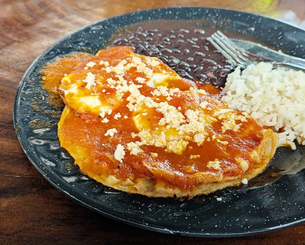 A black metal plate with huevos rancheros for breakfast in Mexico featuring two fried tortillas topped with fried eggs, red sauce, and cheese with black beans and rice on the side.