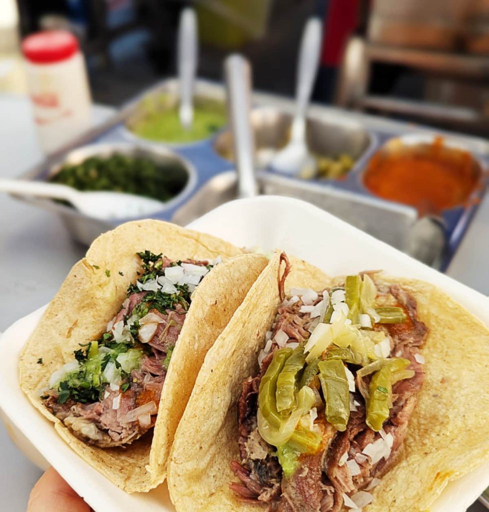 A styrofoam plate with two barbacoa tacos at Los Toños Barbacoa in Mexico City. One is topped with onion and cilantro, the other with long strips of nopal cactus, a tray of salsas blurred in the background.
