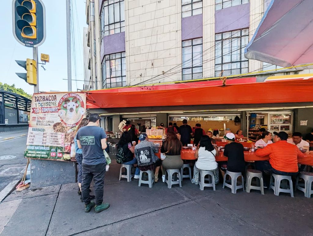 A menu sign reads "Barbacoa y Consomé Los 3 Compadres" beside a red awning shading long tables full of diners seated on stools at one of the best places to eat barbacoa in CDMX.