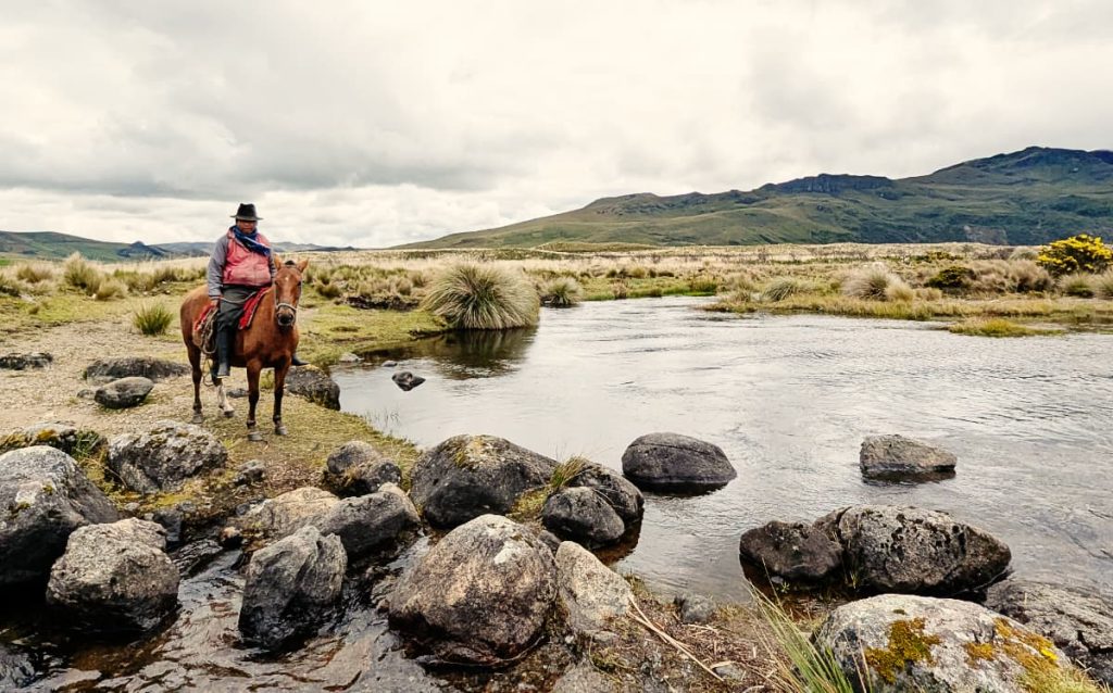 A man on a horse and wearing a cowboy hat next to the lake at Lagunas Ozogoche in Ecuador. In the foreground are large rocks in shallow water. In the background are low mountains and paramo landscapes.