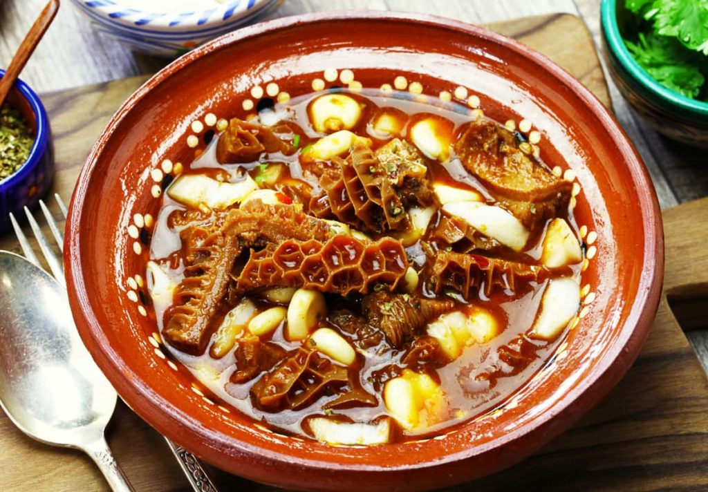 Menudo, a weird food in Mexico, with honeycomb tripe and hominy in red broth, served in a dotted clay bowl with a spoon, fork, dried oregano, and cilantro on the side.
