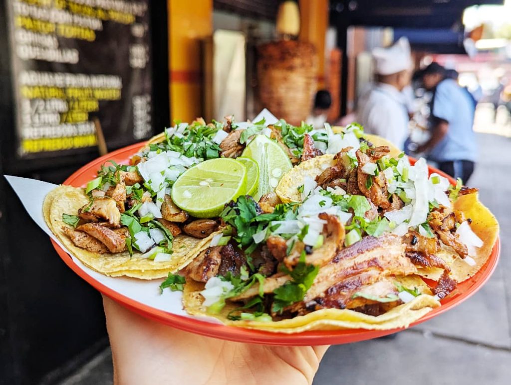 A red plastic plate of five tacos al pastor, one of the most popular Mexican tacos. Each taco is two small corn tortillas piled with fatty pork. On top are several slices of lime. In the blurred background is the trompo with a pineapple on top.