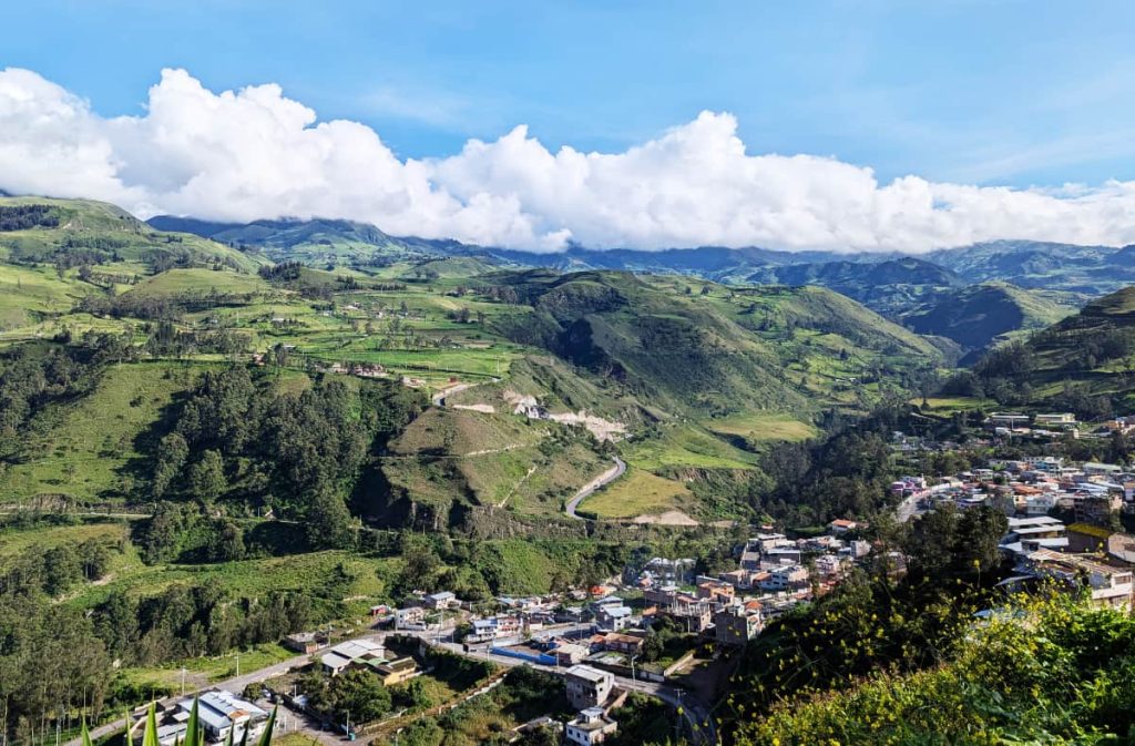 The view from the mirador in Alausí Ecuador featuring bright green mountains with houses in the valley below. Low, puffy white clouds hug the mountaintops under a blue sky.