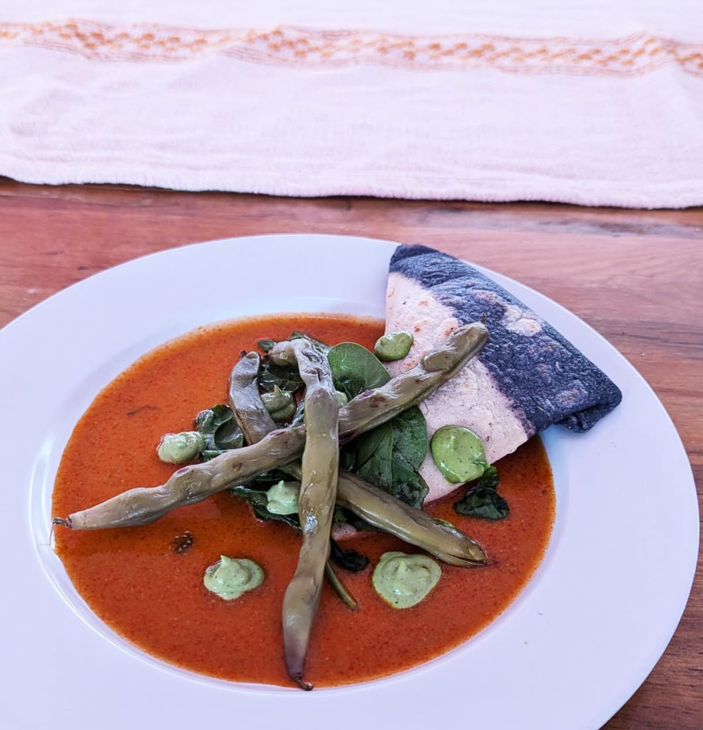 Mole de caderas, a unique Mexican dish, features a red-orange broth with green beans, leafy greens, and dots of green sauce, served with a folded blue and white corn tortilla on a white plate.