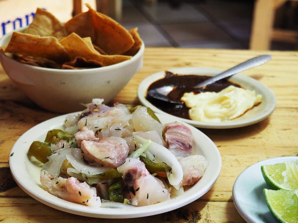 Pickled cow feet, one of the weird foods in Mexico, with onion, jalapeño, and dried herbs on a white plate, served with a bowl of tortilla chips, limes, and a plate with a dark sauce and a creamy white sauce with a spoon.