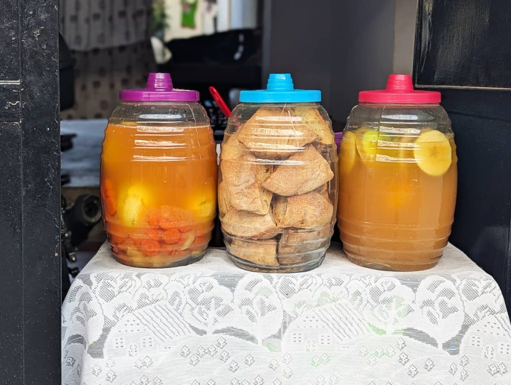 Three plastic barrel jars on a lace tablecloth with ingredients to make piedrazo, one of the unique foods in Mexico. Large pieces of hard bread in the center, pickled carrots, mango, and a golden vinegar in the containers on either side.