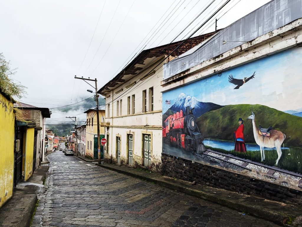 Looking down a cobblestone street in Alausí with a prominent piece of street art featuring a woman followed by a llama next to the train tracks with a red and black train. In the background is a body of water and snow capped mountains with a condor flying overhead. The scene is taken on a cobblestone street in Alausí.
