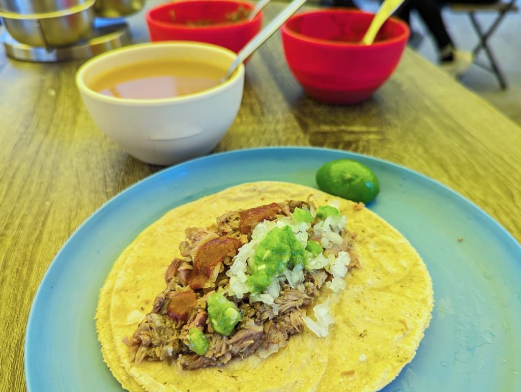 A barbacoa taco topped with onion, red and green salsas sits on a blue plate with a lime wedge, next to a bowl of consomé and two bowls of salsa on a wooden table.