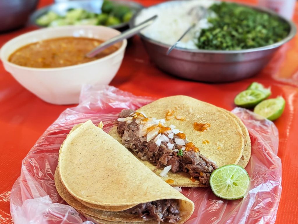 Two tacos de barbacoa in Mexico City topped with onion and red salsa, lime wedges on the side, with a bowl of consomé and a larger bowl of chopped onion and cilantro in the background.