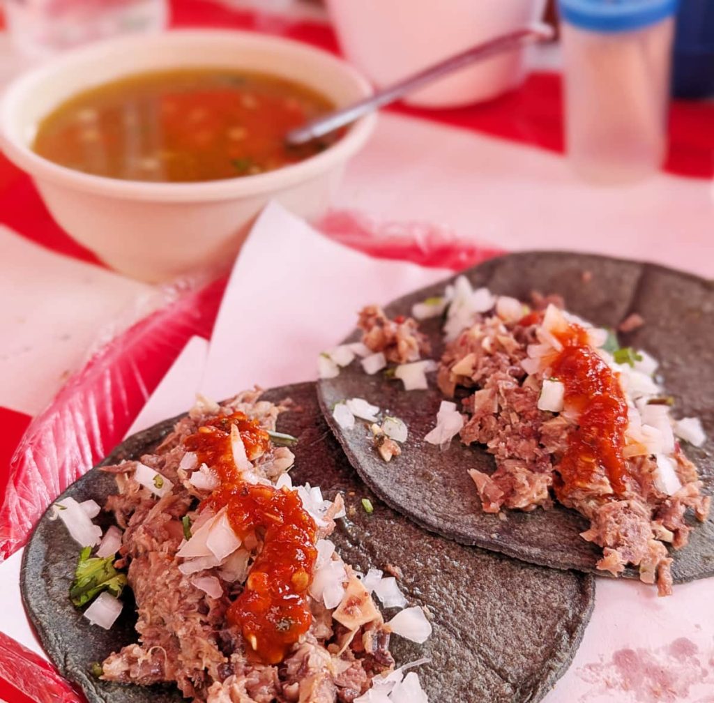 Two barbacoa tacos on blue corn tortillas topped with onion, cilantro, and red salsa, with a bowl of consomé blurred in the background.