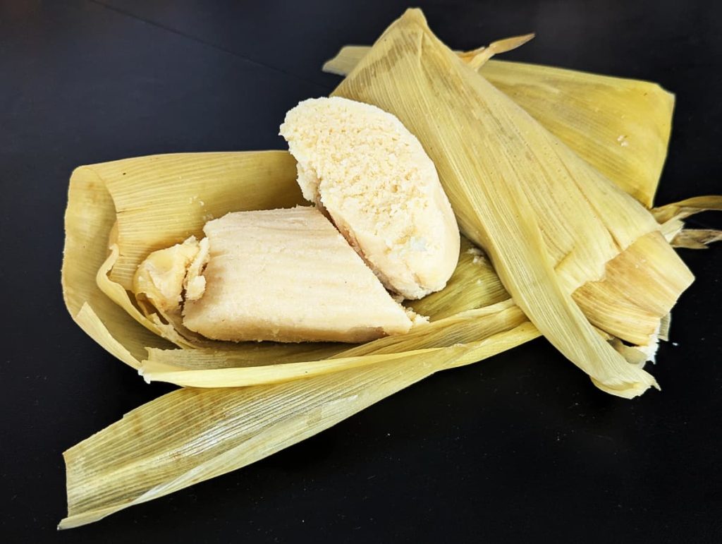 A pale yellow tamal de elote, one of the classic types of sweet tamales, cut in half on open corn husks against a black surface.