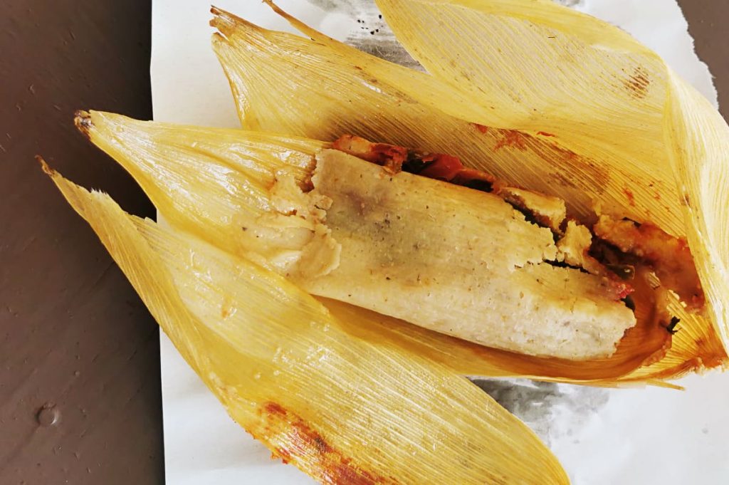 An unwrapped tamal de rajas on yellow corn husks, with red tomato sauce and dark green chile strips peeking out one end, on torn butcher paper.