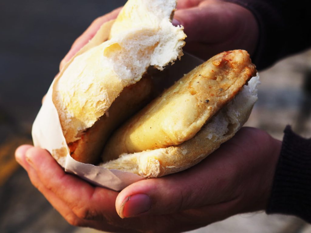 Two hands hold a torta de tamal, a square yellow tamal tucked into a split white bread roll wrapped in butcher paper.