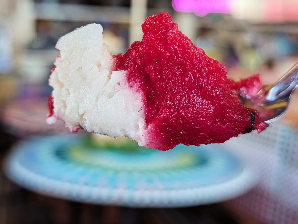 A closeup of a spoonful of white leche quemada and bright red tuna (prickly pear) nieves ice cream, held against a blurred blue table.