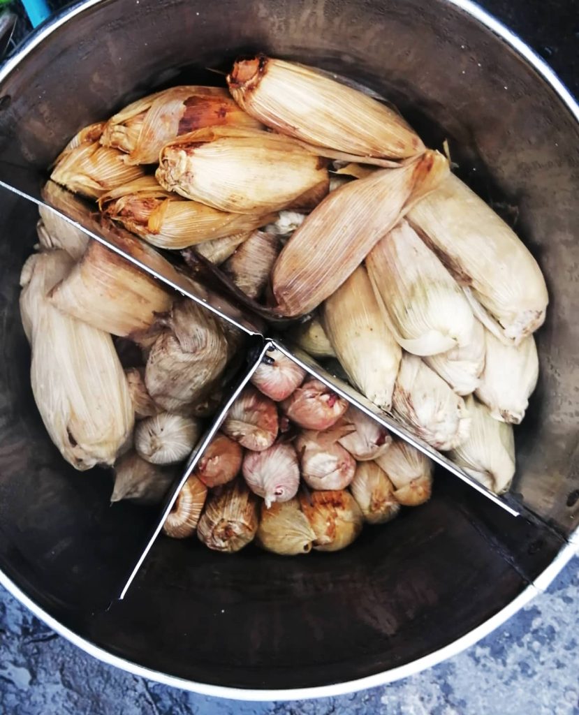 Overhead view of a steel pot split into three sections filled with corn husk tamales, some husks stained red and others tinged pink, sorted by types of Mexican tamales.