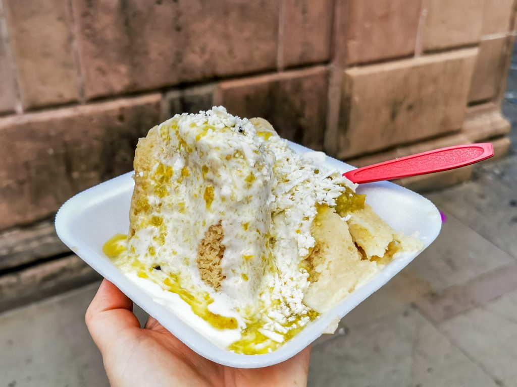 A hand holds a styrofoam tray of uchepos, one of the types of tamales in Mexico, smothered in crema, crumbled cheese, and green salsa, with a red plastic spoon and a pinkish stone wall behind.