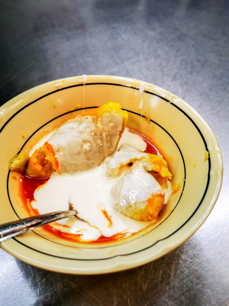 Two uchepos in red salsa drizzled with crema, served in a yellow bowl with black stripes on a stainless steel counter.