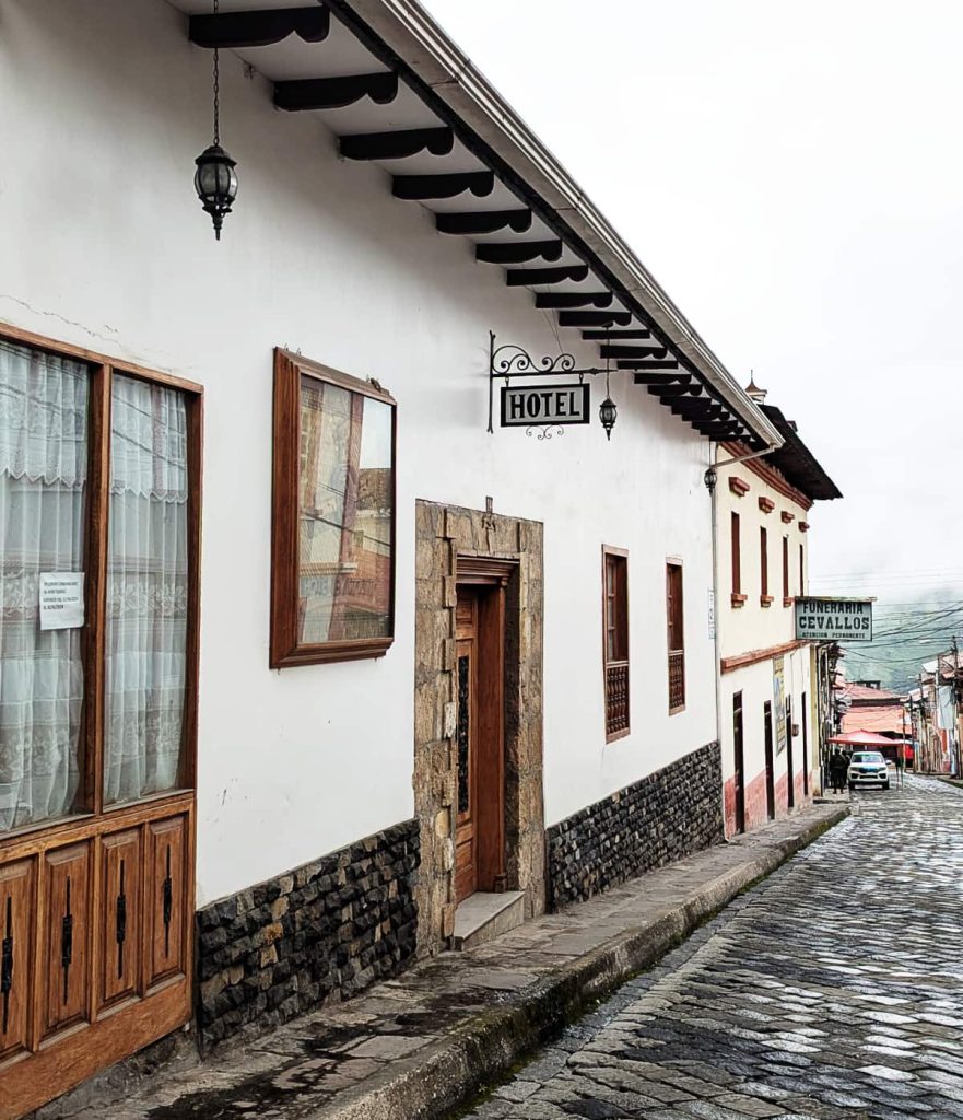 A white building with a wrought-iron hotel sign leans over a quiet cobblestone street in Alausí.