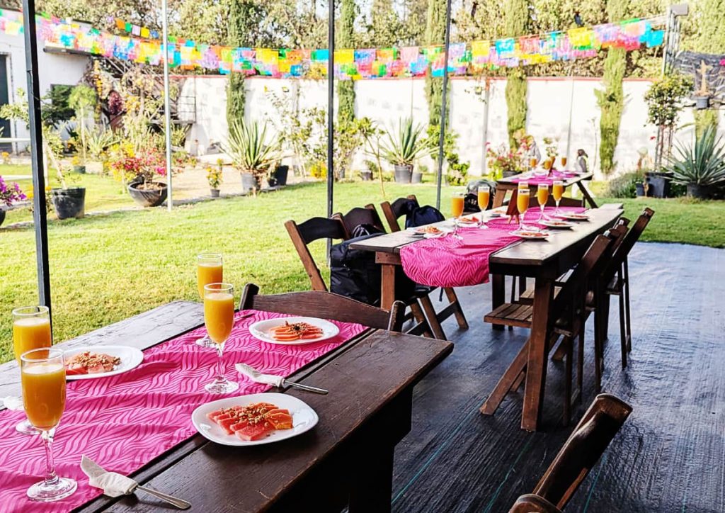 At the end of a Xochimilco kayak tour, sliced papaya topped with seeds on white plates and mimosas in champagne flutes are set on a wooden table with a pink table runner. Strings of colorful papel picado are hanging over the grass in the background.