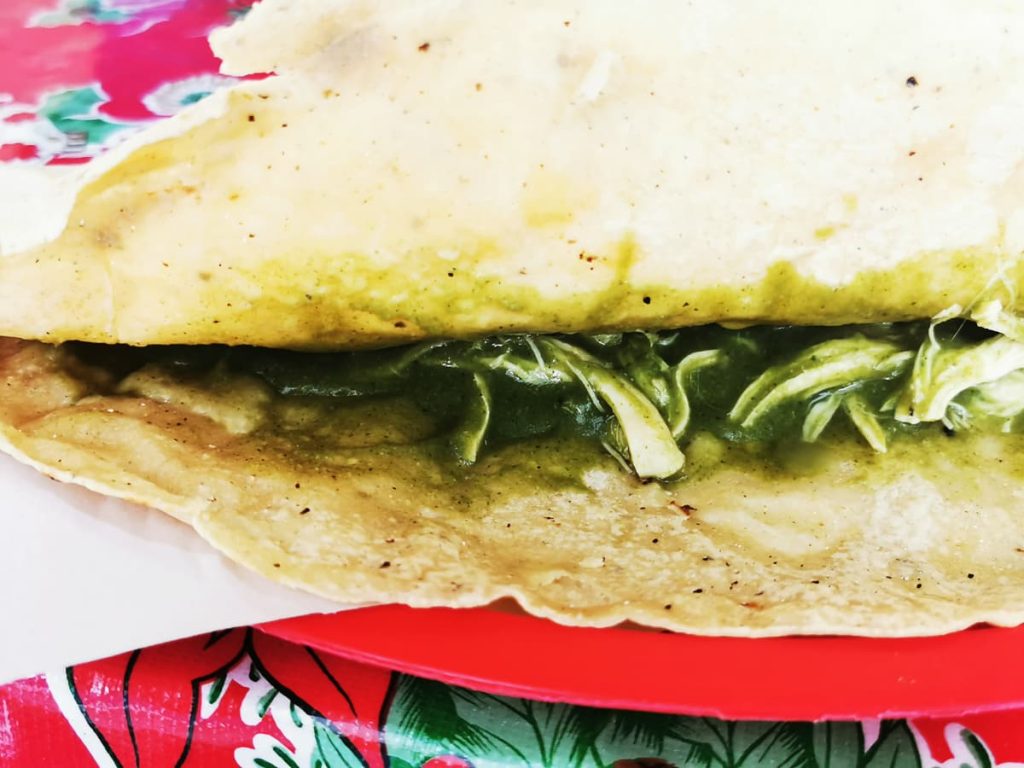 Large empanada folded open to show shredded chicken in green salsa, on white paper and a red plastic plate over a red floral tablecloth.