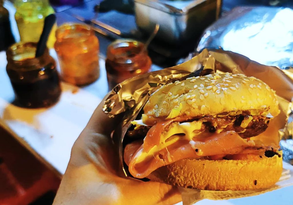 At a street stall in Mexico, a hand holds a hamburger wrapped in gold foil, stacked with a patty, ham, and melted cheese on a sesame bun, with jars of salsa behind.