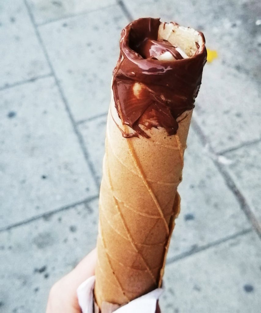 A hand holds a crispy rolled marquesita filled with banana slices and with nutella coating the open end, above a gray paved sidewalk.