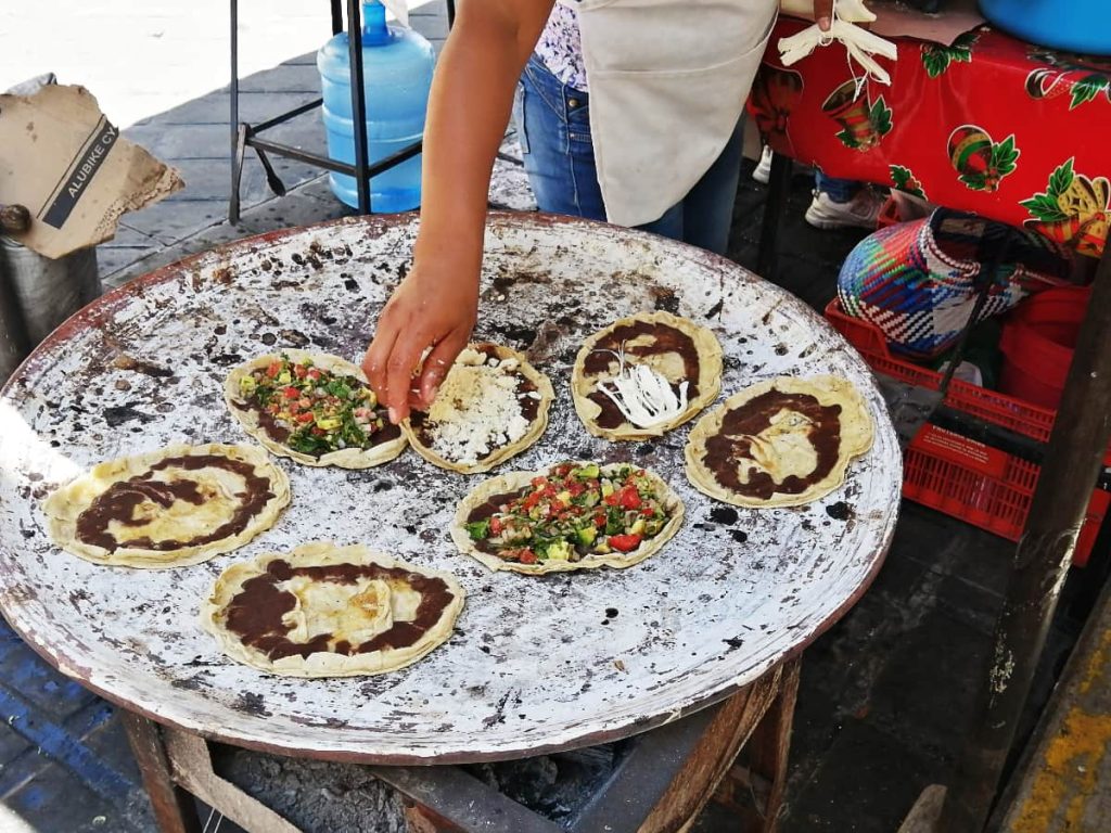 A vendor tops flat, oval memelas on a large comal with black beans, quesillo, crumbled cheese, and chopped tomato and avocado.