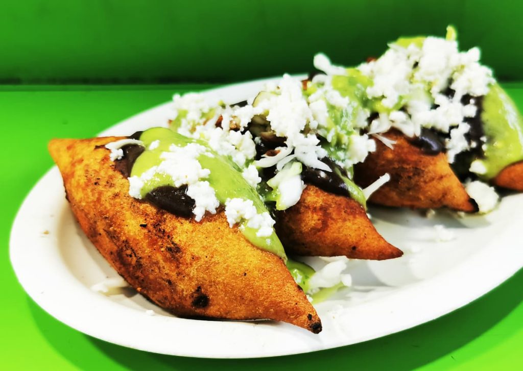 Four fried, pointed masa pockets topped with black bean paste, green salsa, and crumbled white cheese on a white plate.