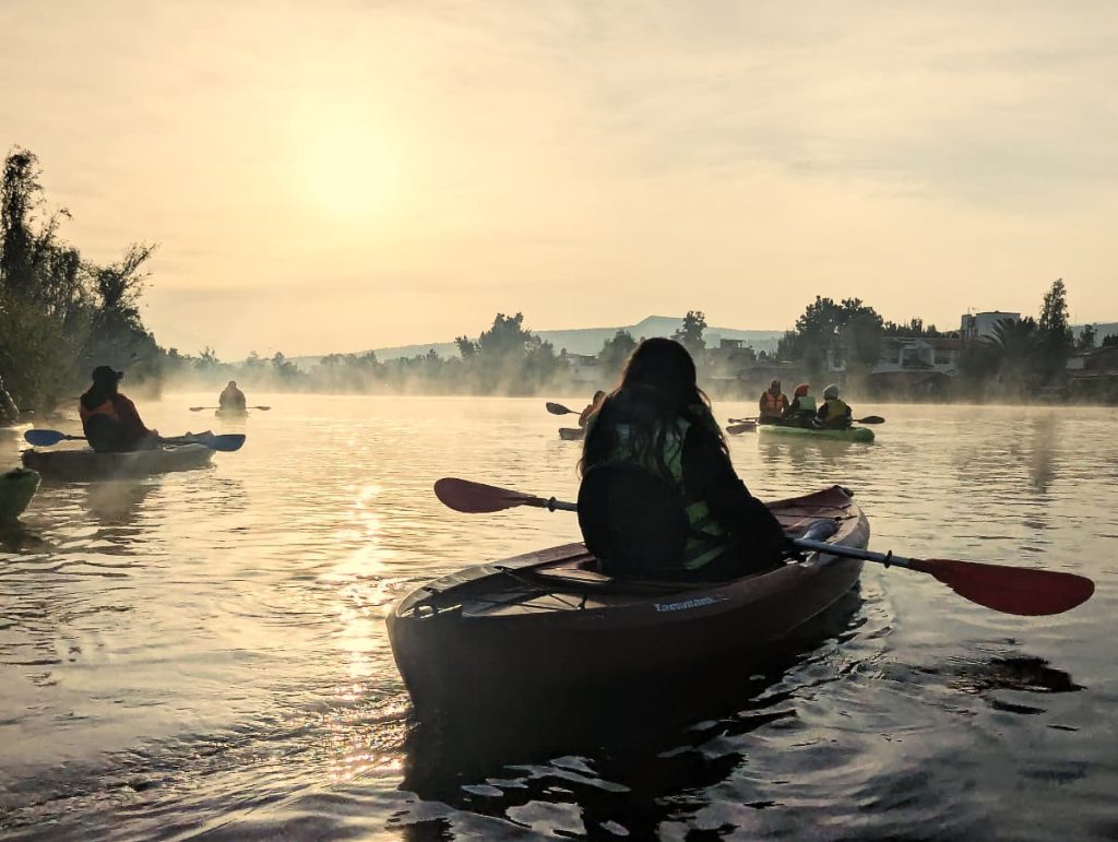 Kayakers paddle across an open stretch of water on a kayak Xochimilco tour, mist rising around them as the sun breaks low over distant hills.