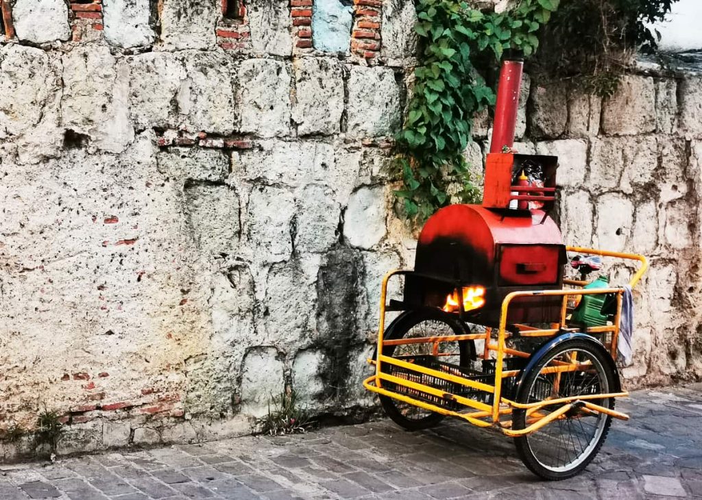 A mobile street food cart in Mexico with a red metal oven, tall chimney, and glowing flame sits on a yellow three-wheeled tricycle against a weathered stone wall with ivy.