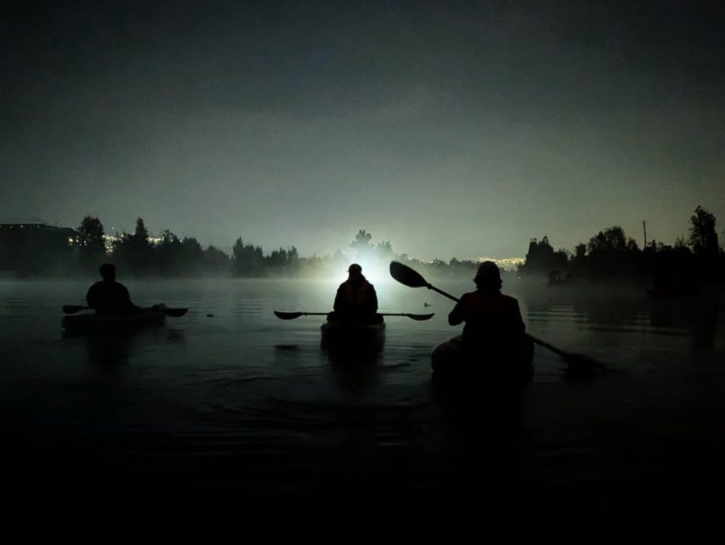 Silhouette of three kayakers paddling in near-darkness at the start of a sunrise kayak in Xochimilco, city lights glowing through the mist on the horizon.