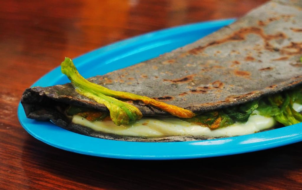 A blue corn quesadilla filled with melted white cheese and orange squash blossoms on a blue plastic plate.