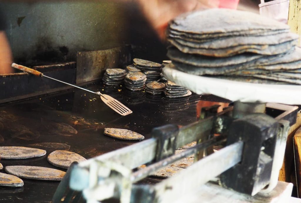 A flat griddle with blue corn tlacoyos cooking in the foreground and a metal spatula resting on the edge. Stacks of tlacoyos are resting in the back and a stack of blue corn tortillas are on a scale on the right.