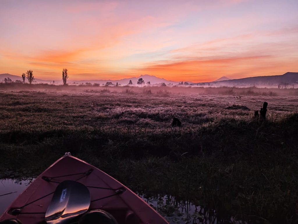 The bow of a kayak in Xochimilco rests at the edge of a frost-covered field, with an orange sunrise sky and a volcano’s silhouette on the horizon.