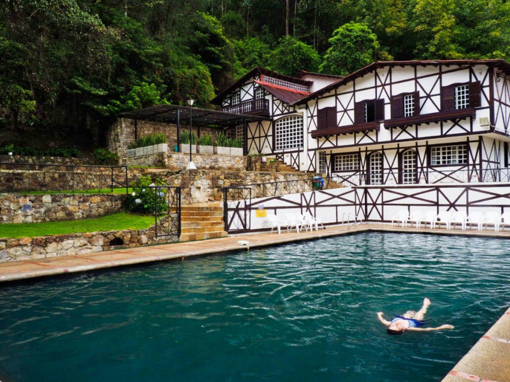 A woman floats on her back in the thermal pool at termales los volcanes. Behind the pool is the white hotel building with wooden accents.