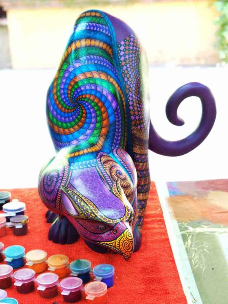 A colorful alebrije that resembles a mix of a panther and a large bird sits on a table in front of a palette of paints.