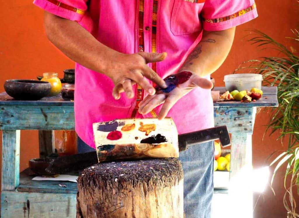 On an alebrije tour, a man spreads different colors of natural paints on his hand to demonstrate how the colors are developed.