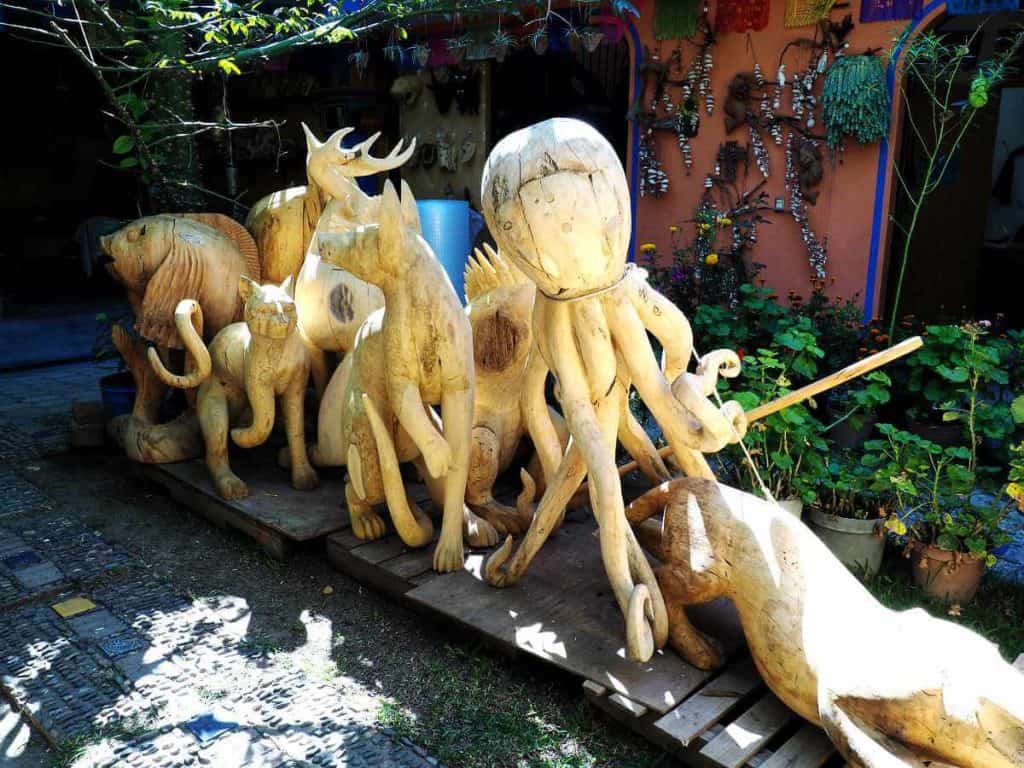 Larged carved alebrijes sit on a wooden palette in the sun while they dry. Figures include a large octopus, several mammals, and fish.