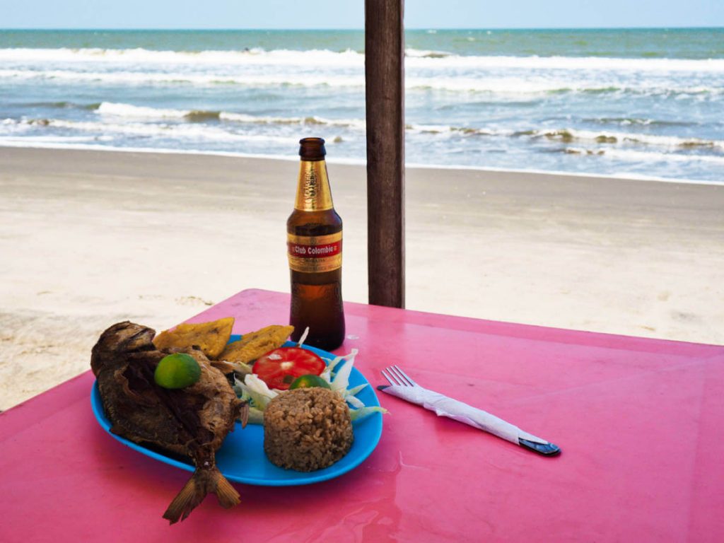 At one of the beaches in Cartagena, La Boquilla, a Club Colombia beer bottle sits with a plate of fried whole fish and coconut rice on a table over looking the ocean.