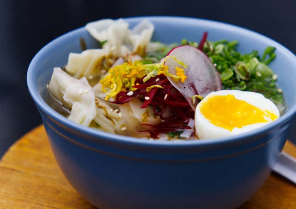 A blue bowl of Ramen noodles with dumplings topped with sliced radish, shredded beets, and squash blossoms. A bright yellow poached egg sits open face on the edge of the bowl.