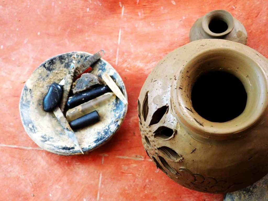 A bird's eye view of a raw Oaxacan black pottery vessel decorated with a flower cut into the side. Next to it, potter's tools lay in a bowl on the floor.