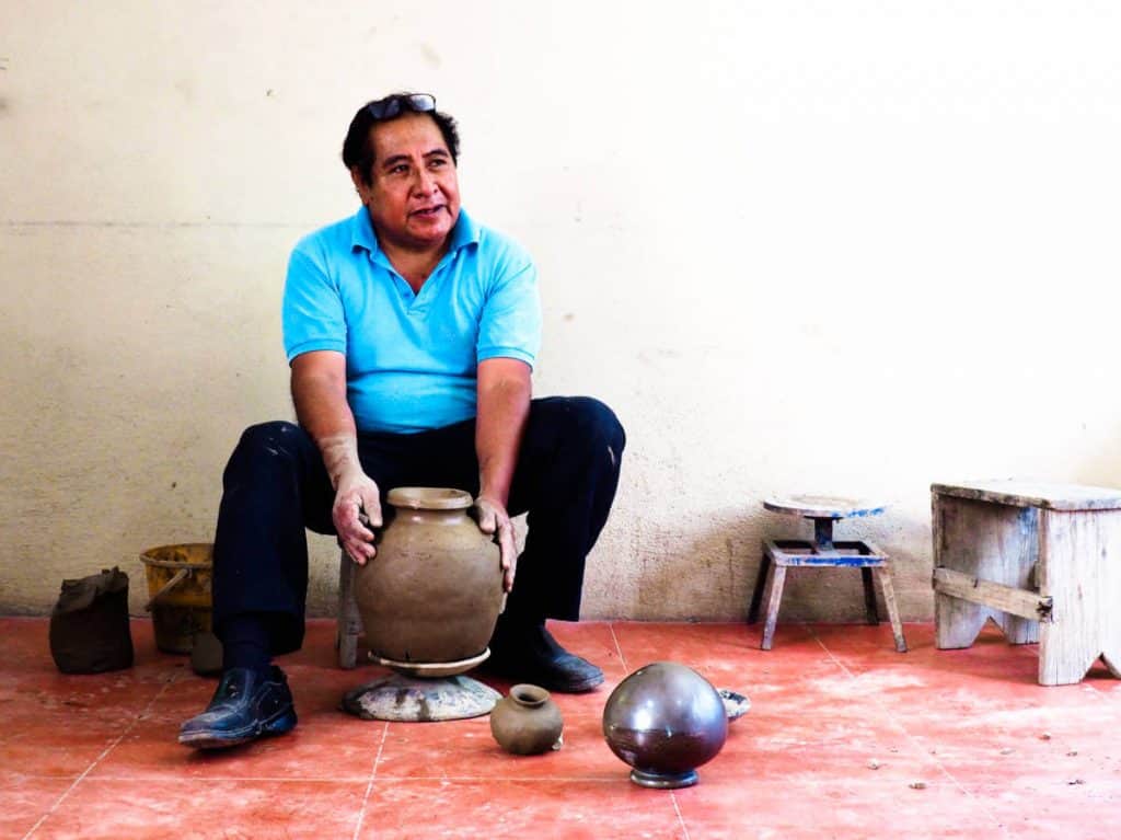 Sitting on a low stool, Fabian makes the finishing details on a clay vessel that sits on a short platform on the ground.