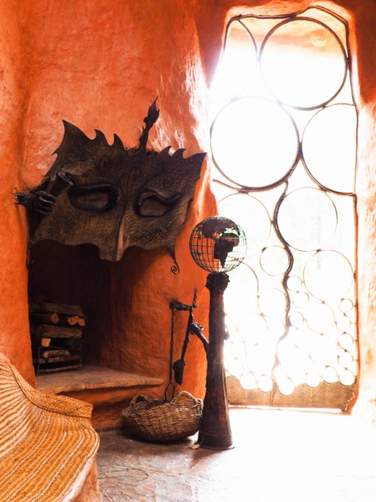 A large metal mask frames the top of the main fireplace which is made of clay. To the right of the fireplace is a door to the outside created of metal circles in various sizes.