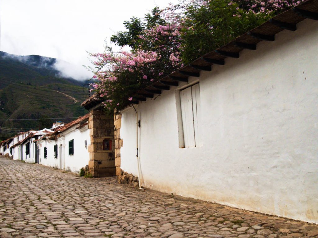 Pink bougainvillea flowers grow over the top of the wall, one of many white colonial houses lining the cobblestone streets of Villa de Leyva. Fog settles over the mountains in the background.