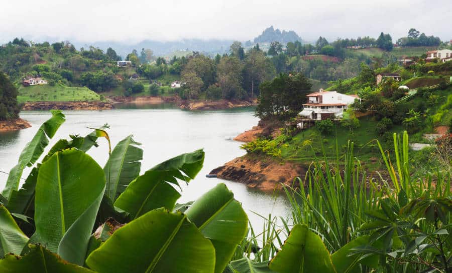 A view near Galeria Guatape Hostel of the Guatape lake where you can see a prominent house overlooking the lake andtropical banana trees in the foreground.