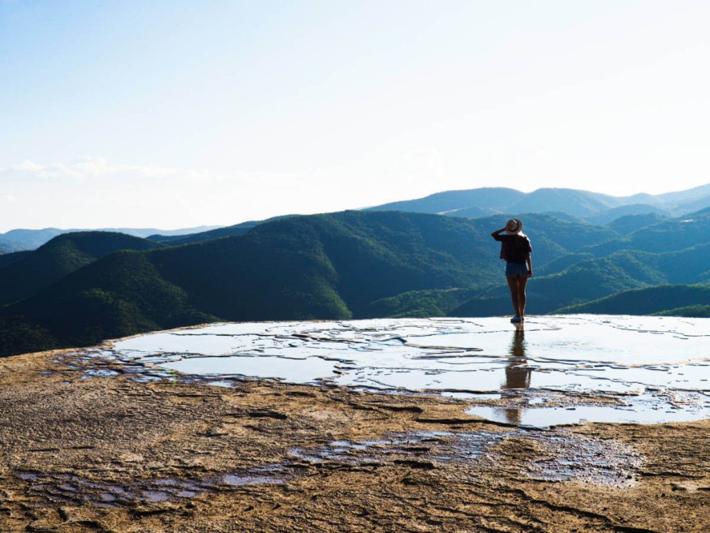 A woman holds on to her hat as she stands at the edge of the cliff, overlooking the mountains and valley at Hierve el Agua