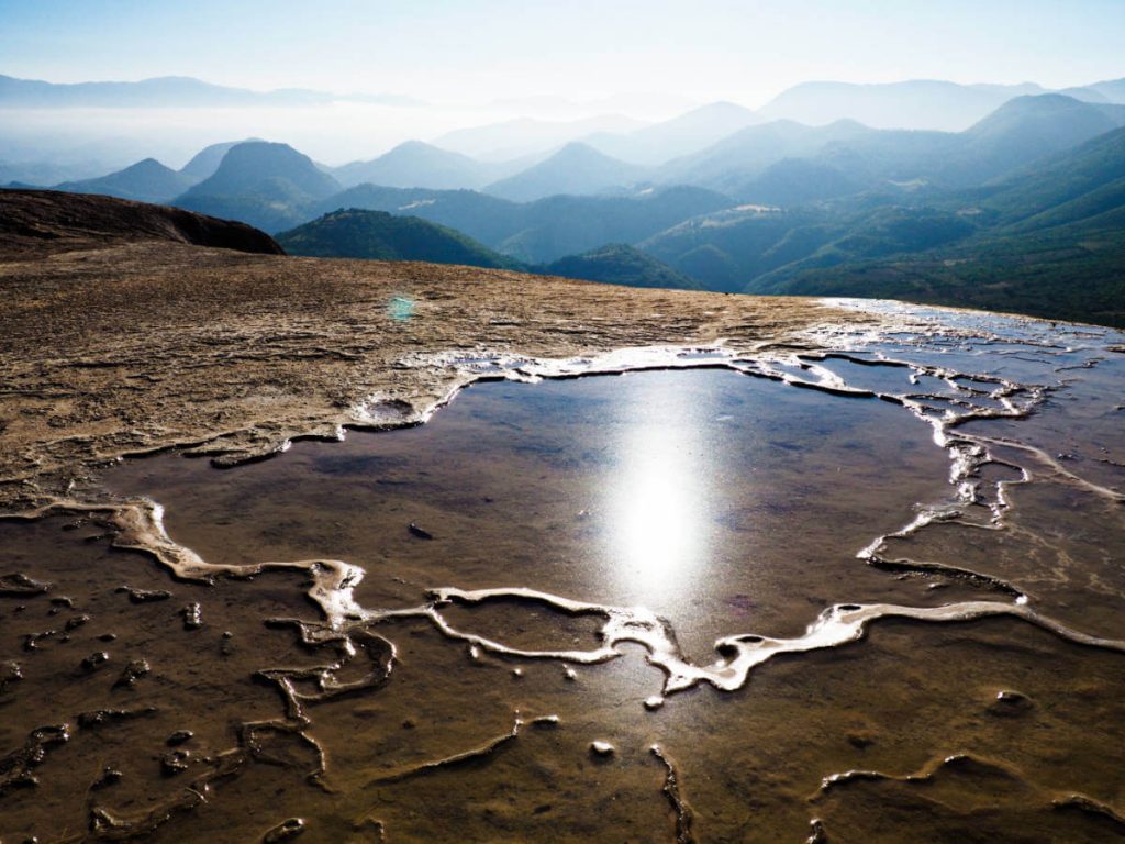 The sun reflects in a shallow pool of water along the trail at Hierve el Agua with mountaintops in the background.