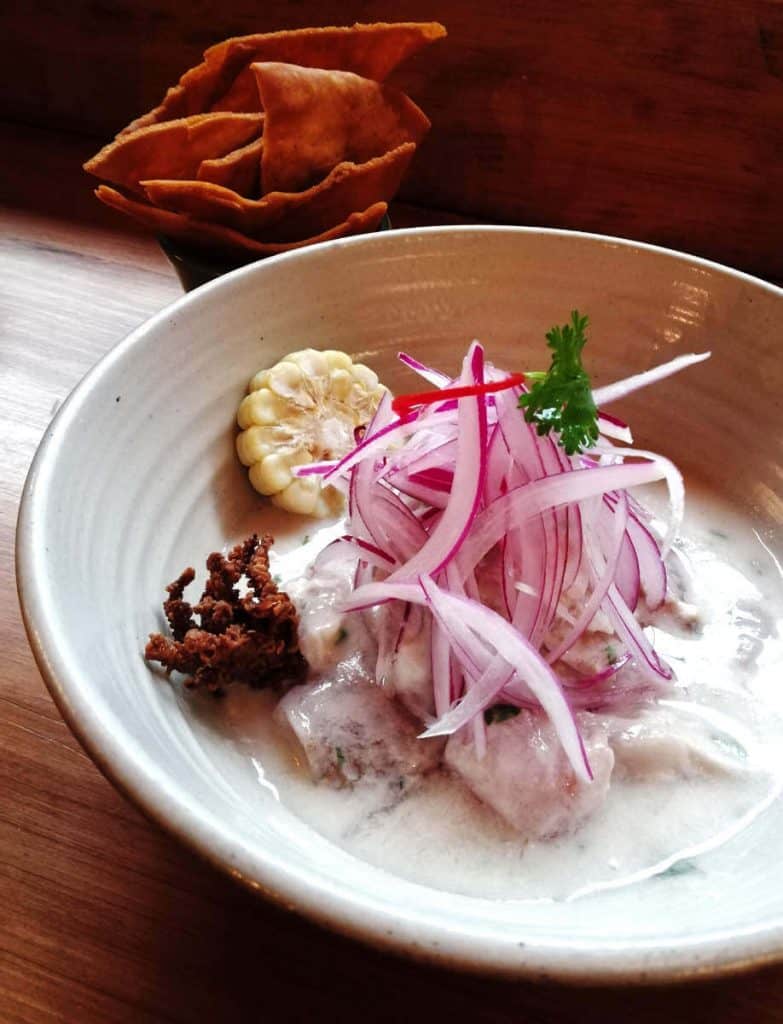 A bowl of Peruvian ceviche, smothered in a leche de tigre sauce and topped with slivers of raw red onion. Plated with a small crispy calamari and thin slice of corn on the cob.