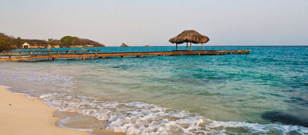 Small blue ocean waves come ashore the off white sand at Playa Libre on Isla Grande, Colombia with a long dock and palapa in the background.